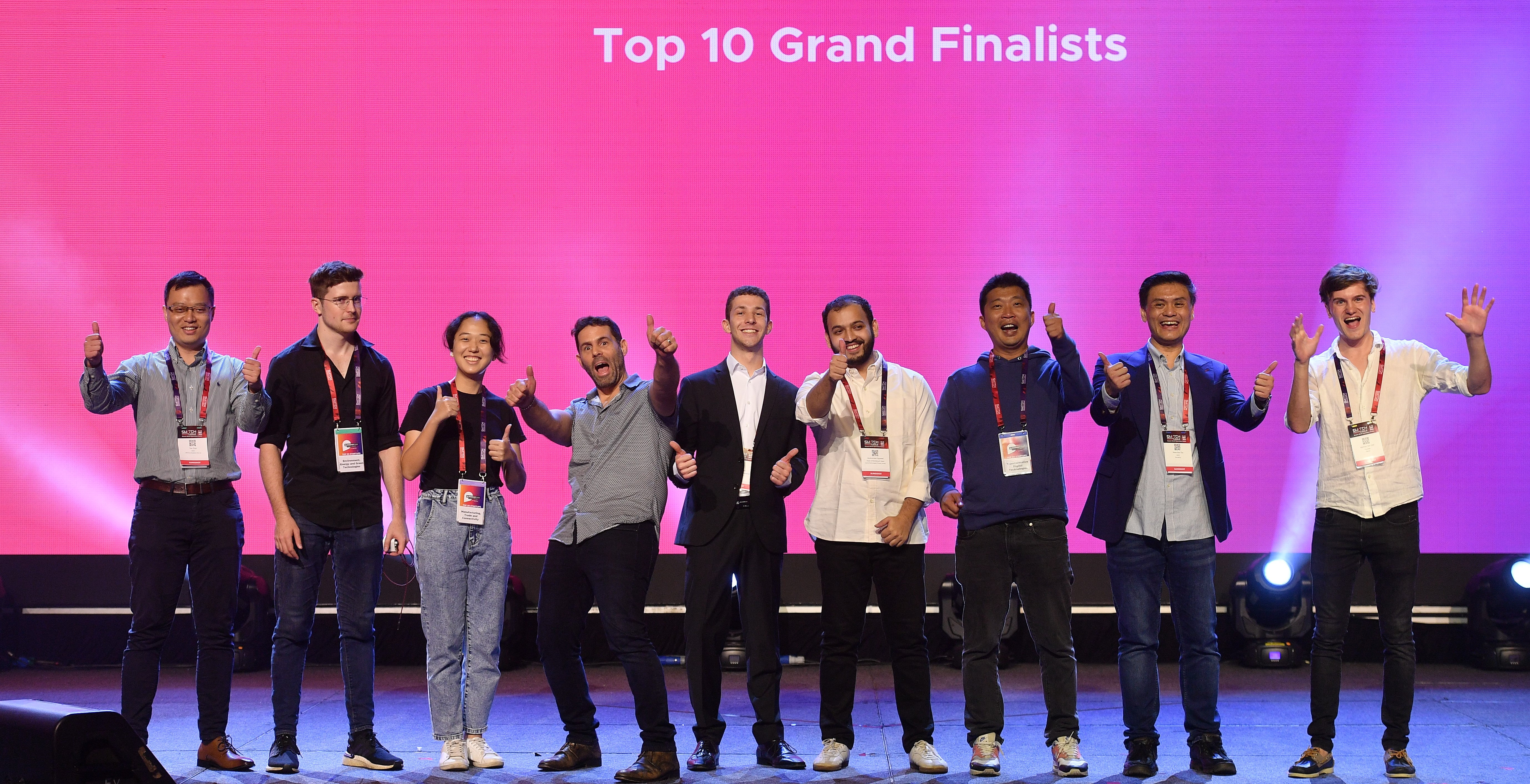 Nine people pose with thumbs up in front of a pink background, wearing name tags and lanyards.